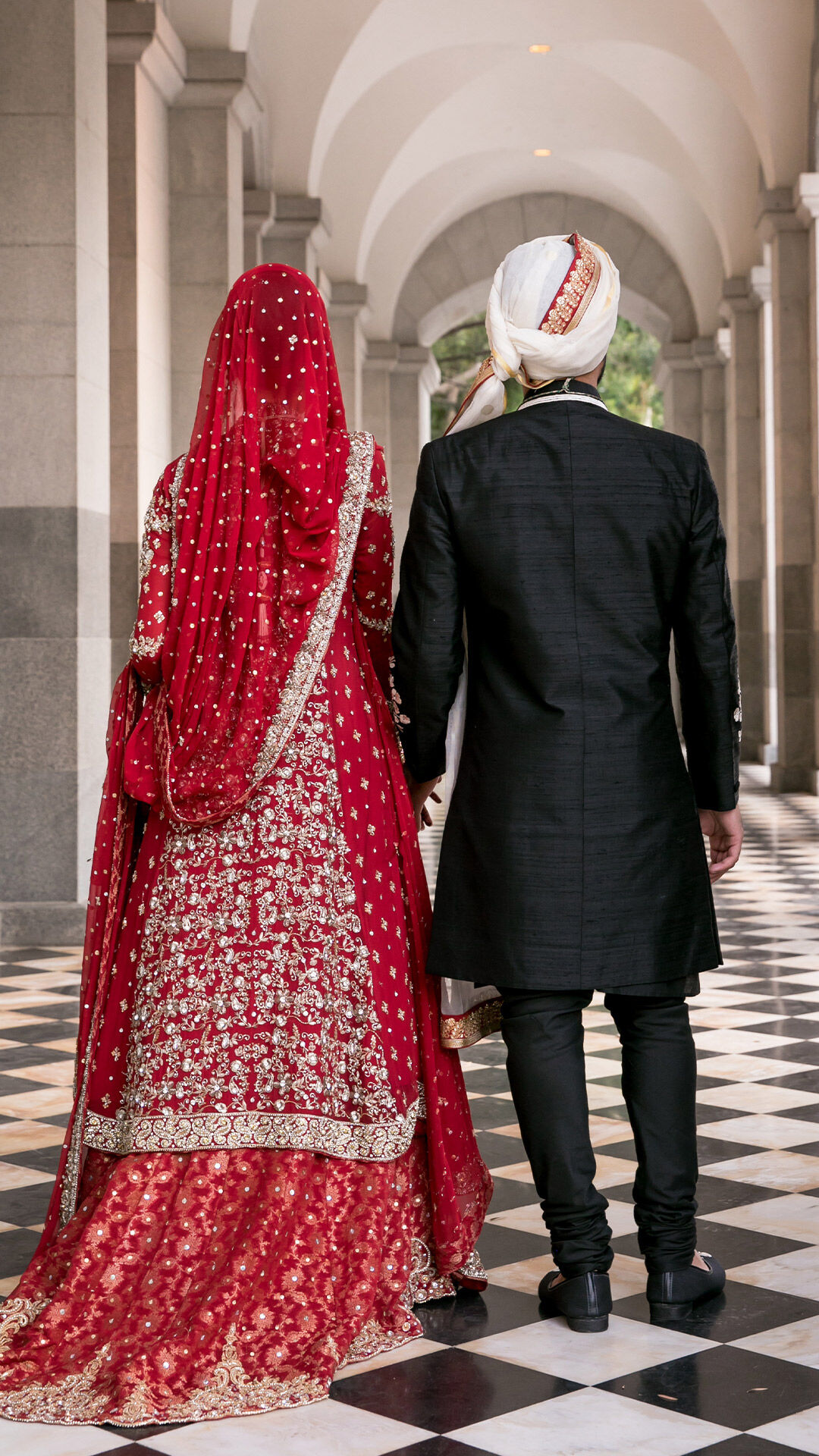 Punjabi bride and groom in traditional wedding attire during Roka ceremony, walking hand-in-hand in a grand hallway.